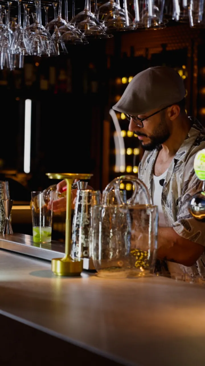 A bartender in a flat cap mixes a drink at the VÁ BAR counter, with glasses hanging above.